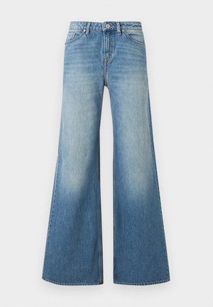 Žena oblečená v hnedom saku, béžovom roláku, širokých modrých džínsach, bielych podpätkoch a slnečných okuliaroch, stojaca pred jednoduchým svetlým pozadím.