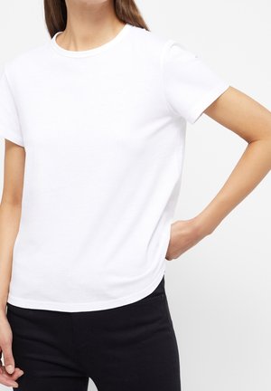 Woman wearing a plain white short-sleeve T-shirt and black pants, with one hand on her hip and the other relaxed by her side.