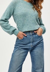 Maglione azzurro chiaro con texture, maniche lunghe e polsini a costine, indossato con jeans in denim blu a vita alta e gamba dritta.