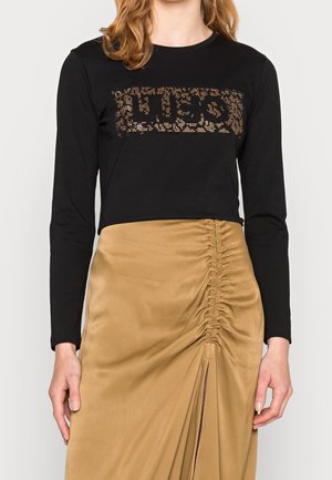 Woman wearing a black long-sleeve top with a studded leopard print logo and a tan ruched skirt with a front slit.