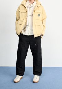 Giacca gialla con molte tasche, chiusura con zip, indossata sopra una camicia bianca a fantasia, abbinata a pantaloni neri e sneakers bianche con suola marrone.