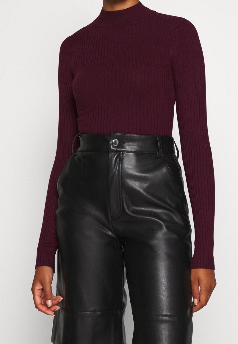 Haut col roulé côtelé bordeaux associé à un pantalon en cuir noir taille haute, présentant une texture lisse et des détails à boutons.