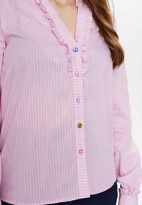 Blusa a righe rosa con scollatura arricciata, impreziosita da bottoni colorati: blu, rosa, viola e verde. Maniche lunghe con polsini arricciati.