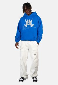 Blue hoodie with a graphic of two white birds, paired with white cargo pants featuring pockets and black accents, worn with black and white sneakers.