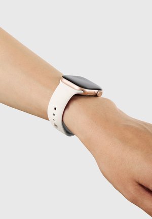 Handgelenk mit einer Smartwatch mit goldenem Rahmen, schwarzem Bildschirm und weißem Sportarmband vor hellem grauem Hintergrund.