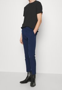 Man som bär en svart kortärmad skjorta, åtsittande marinblå byxor och svarta läderankelkängor, står med händerna i fickorna.