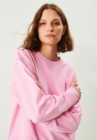 Ung kvinna med axellångt brunt hår bär en rosa sweatshirt, armarna korsade, tittar rakt in i kameran med ett neutralt uttryck.