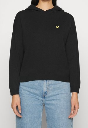 Nainen, jolla on musta huppari, jossa on pieni keltainen lintulogo rinnassa, ja vaaleansiniset farkut.