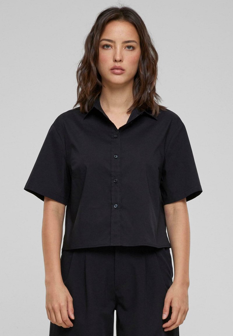 Camicia nera cropped con bottoni, colletto, maniche corte e con una texture di tessuto liscia. Presenta un orlo arrotondato e bottoni abbinati.