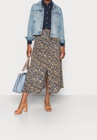 Jeansjacke, marinefarbene Hemd, floralen Midirock in Marineblau, Gelb und Weiß, hellbraune Blockabsatzsandalen und hellblaue Handtasche.
