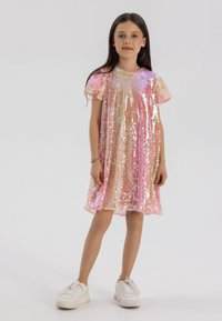Robe rose brillante ornée de sequins multicolores, avec des manches volantes, une coupe ample et une longueur au genou. Portée avec des baskets blanches.