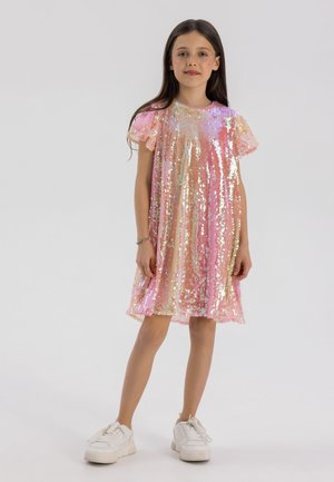 Robe rose brillante ornée de sequins multicolores, avec des manches volantes, une coupe ample et une longueur au genou. Portée avec des baskets blanches.
