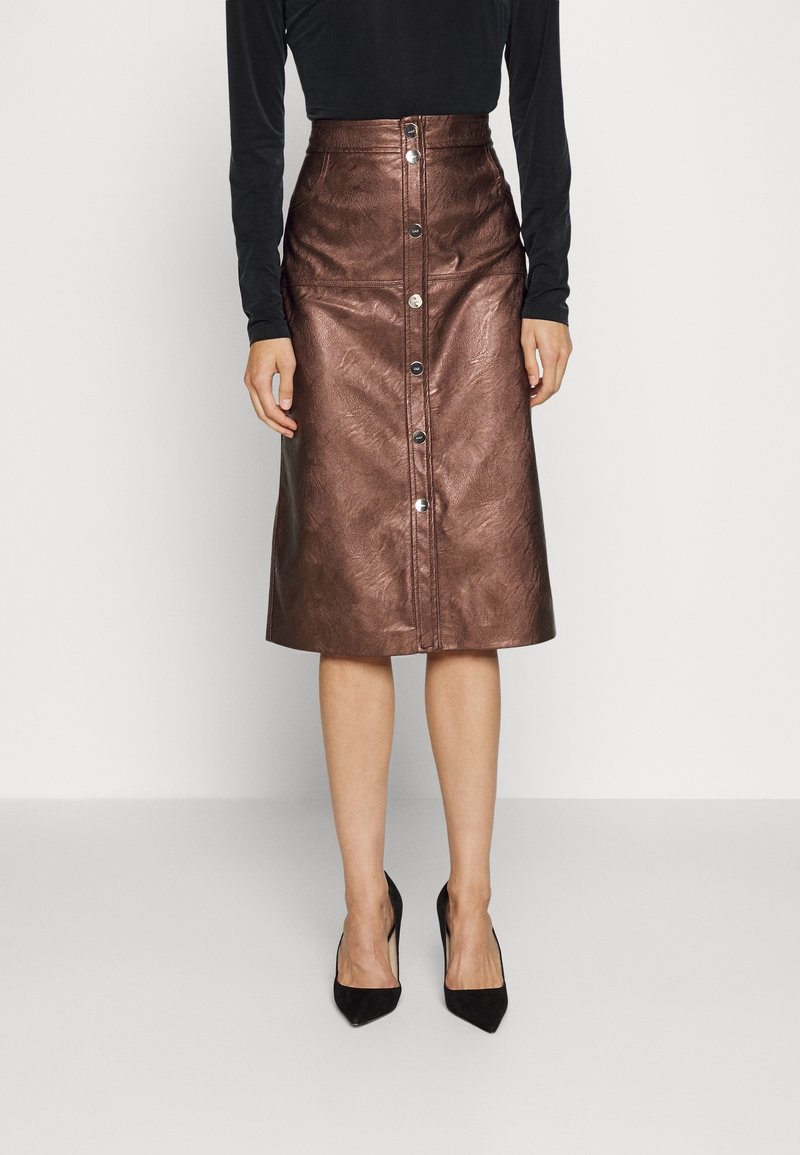 Femme portant une jupe en cuir marron mi-longue avec des boutons sur le devant, un haut noir à manches longues et des escarpins noirs pointus.