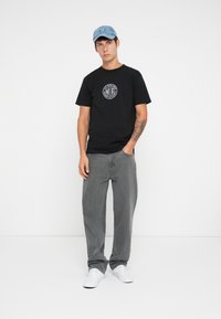 Black short-sleeve T-shirt with a circular graphic design, paired with loose-fitting gray jeans and white sneakers; denim cap completes the look.