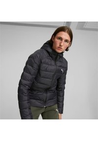 Giacca a vento nera con cappuccio, dotata di chiusura con zip e cuciture orizzontali visibili. Accompagnata da pantaloni verde oliva.