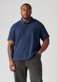 Marineblauwe polo met een zachte textuur en korte mouwen, voorzien van een klein rood logo. Draagt in combinatie met donkergrijze broek, wat een ontspannen pasvorm laat zien.