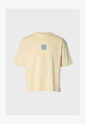 Bézs rövid ujjú póló, amelyen a mellkason középen egy kis kék négyzet alakú logó található, „Grand Amour Hotel” felirattal.