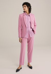 Růžový blejzr a kalhoty z hladké tkaniny, nošené přes růžovou a bílou pruhovanou košili. Černé ploché boty doplňují celý outfit.
