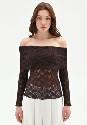 Giovane donna che indossa una maglia nera di pizzo con spalle scoperte e maniche lunghe, abbinata a pantaloni bianchi a vita alta e plissettati, su sfondo semplice.