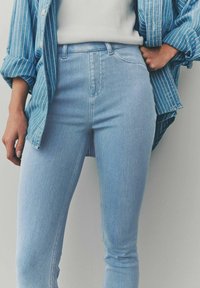 Jeanși skinny bleu deschis cu talie înaltă, cu o textură netedă, asociați cu o cămașă albastră cu dungi, care pune în evidență un croi lejer și o siluetă ajustată.