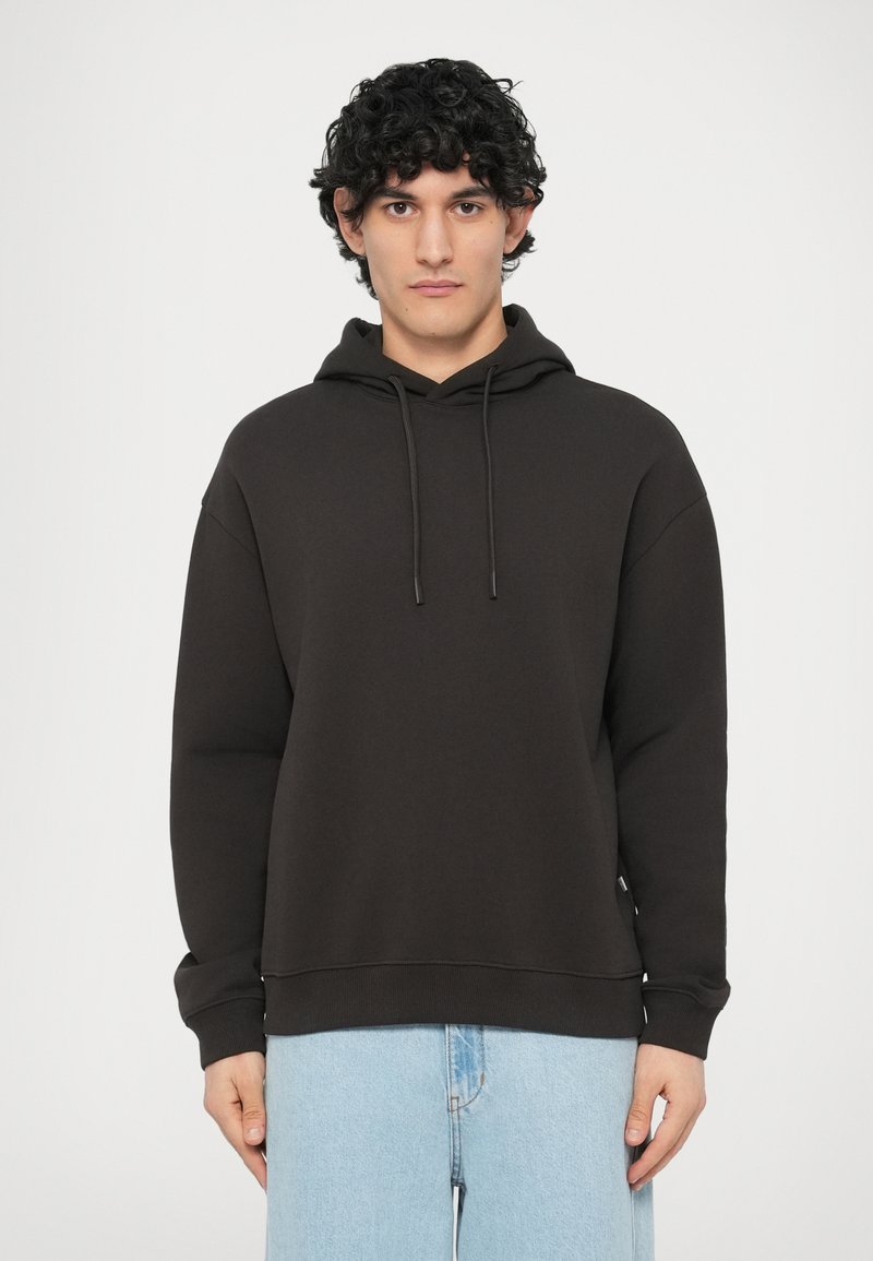 Μαύρο φούτερ με κουκούλα και κορδόνι, άνετη εφαρμογή, ριμπ μανσέτες και στρίφωμα. Συνδυασμένο με ανοιχτόχρωμο μπλε τζιν, σε ουδέτερο φόντο.