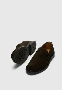 Mocassins en daim marron foncé avec une finition lisse, semelle en caoutchouc comportant des rainures circulaires, et couture décorative à l'avant.