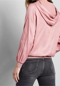 Rosa Kapuzenpullover mit glatter Textur, Ballonärmeln und gerippten Bündchen, kombiniert mit dunkelgrauen engen Jeans.