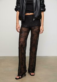 Zwarte doorzichtige kantbroeken met bloemenborduursel en pailletten, strakker bij de taille, wijd uitlopend ontwerp, gecombineerd met een zwarte crop top en jack.