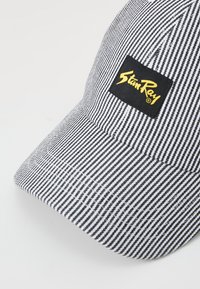 Striped black and white baseball cap with a curved brim. Features a square black patch with yellow logo text sewn on the front.