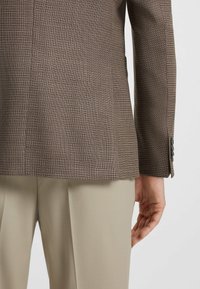 Brun mönstrad kavaj i texturerad tyg, med en sidoöppning och svarta knappdetaljer på ärmarna, i kombination med beige byxor.