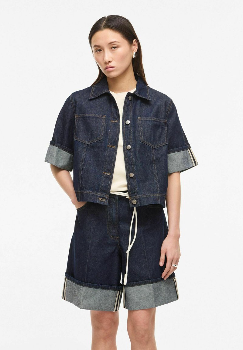 Femeie purtând o geacă din denim închis la culoare cu mâneci scurte rulate și pantaloni scurți evazați din denim, asortați, cu tivuri rulate.