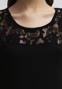 Närbild av en kvinnas övre bröst som bär en svart topp med rund halsringning och blommig spetsöverdrag på den övre delen.