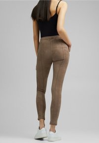 Slim-fit bruine suède broeken gedragen door een vrouw met een zwart mouwloos shirt en witte sneakers, staand tegen een effen grijze achtergrond.