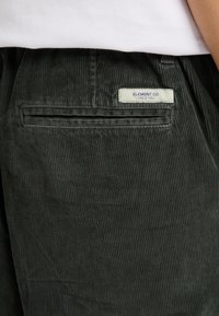 Ciemnozielone spodnie sztruksowe z tylną kieszenią, na której znajduje się etykieta z napisem "ELEMENT CO. SINCE 1992" w niebieskim kolorze. Teksturowany materiał w prążki.