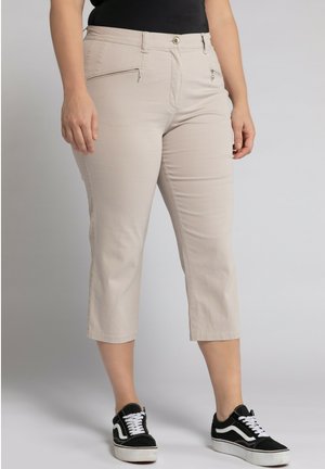 Pantaloni cropped beige con due tasche con zip, un bottone frontale e una texture liscia, abbinati a sneakers nere e bianche.