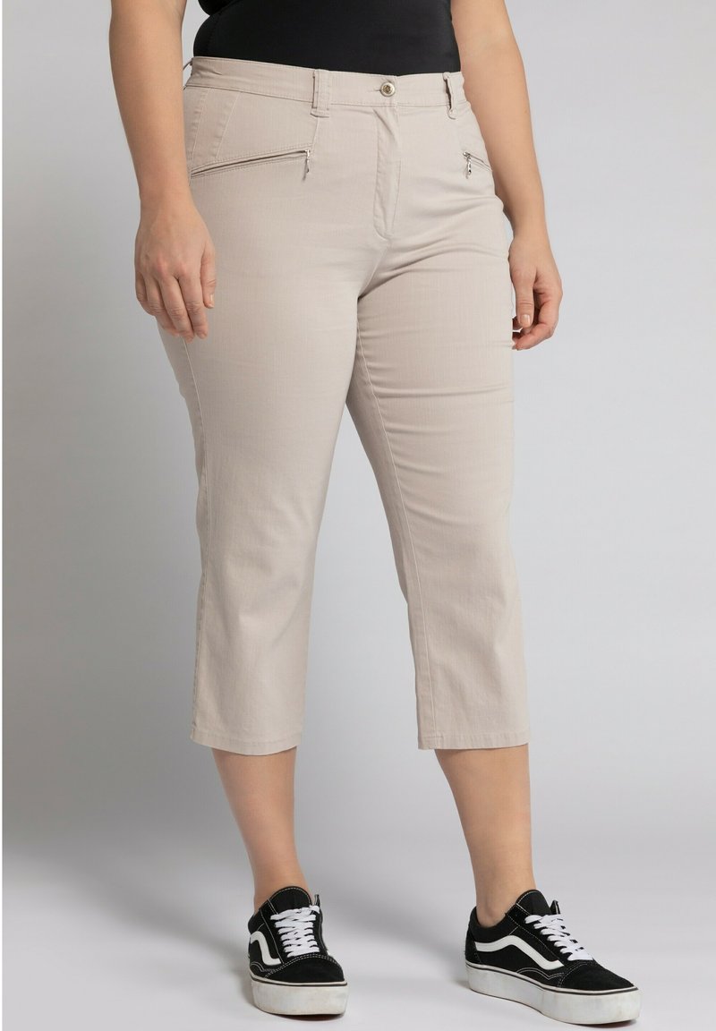 Pantalons beiges coupe courte avec deux poches zippées, un bouton frontal et une texture lisse, associés à des baskets noires et blanches.
