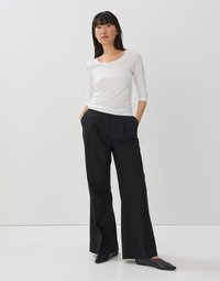 Witte, getailleerde langemouwtop met een ronde hals; gecombineerd met hoge, wijde zwarte broek; model draagt zwarte puntige schoenen.