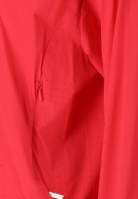 Red jacket with a smooth exterior, featuring a zippered pocket and perforated detailing on the inner lining. Lightweight and breathable fabric.