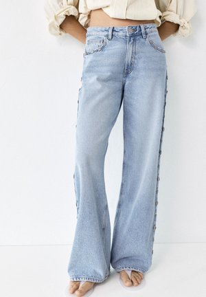 Jean bleu clair à jambes larges avec boutons sur le côté, porté pieds nus, associé à un top beige aux manches retroussées, court à la taille.