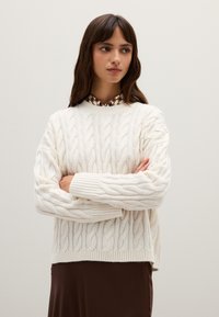 Cremfarbener Kabelstrickpullover mit dicken Rippbündchen und -saum, �über einer blusentypischen braunen gemusterten Bluse getragen. Geschmeidige Textur und lockere Passform.