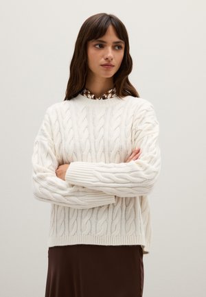 Suéter de punto trenzado color crema con gruesos puños y dobladillo acanalados, llevado sobre una blusa marrón con patrón y cuello. Textura suave y corte relajado.