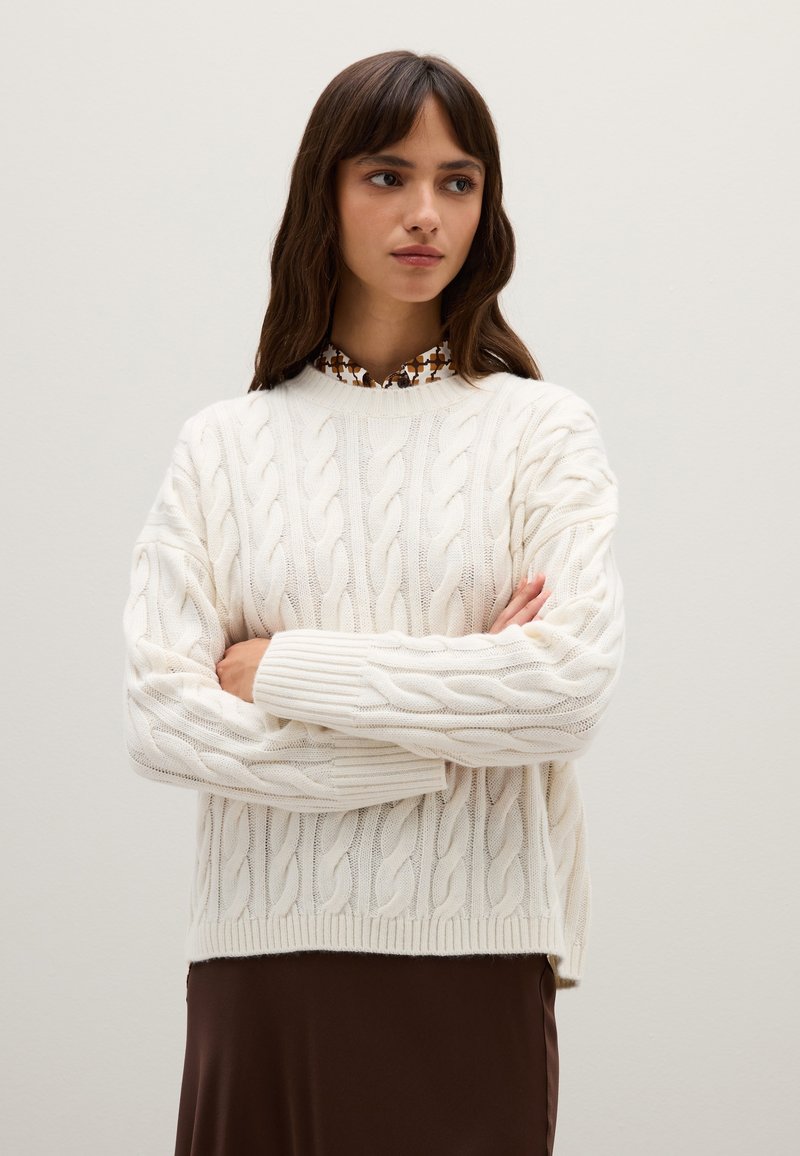 Cremfarbener Kabelstrickpullover mit dicken Rippbündchen und -saum, über einer blusentypischen braunen gemusterten Bluse getragen. Geschmeidige Textur und lockere Passform.