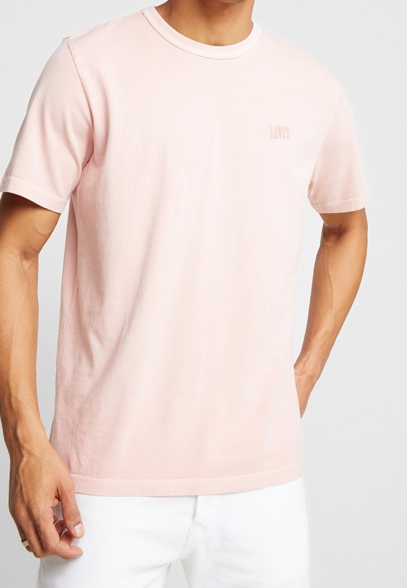 T-shirt en coton rose clair à manches courtes, col rond et détail de logo subtil. Texture lisse, coupe régulière, associé à un pantalon blanc.