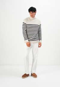 Giovane uomo che indossa un maglione a collo alto a righe crema e navy, pantaloni bianchi e scarpe slip-on marroni, in piedi contro uno sfondo bianco.
