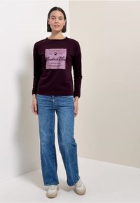 Frau trägt einen dunkelvioletten Sweatshirt mit dem Text "Mountain Moon", blaue Jeans und weiße Sneakers, steht vor einer zweifarbigen beigefarbenen Wand.