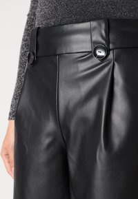 Pantaloni in pelle nera con una texture liscia, caratterizzati da una vita alta, pieghe frontali e due grandi bottoni rotondi.
