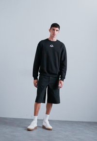 Svart sweatshirt med en liten broderad design, kombinerad med svarta shorts. Modellen bär vita sneakers och sportiga vita ankelstrumpor.