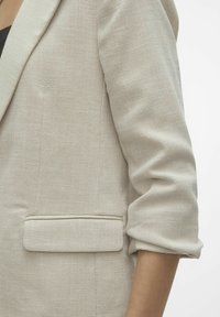 Beige texturerad kavaj med uppvikta ärmar och en enda ficka fram. Tyget har ett subtilt mönster och en strukturerad design.
