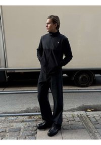 Młody mężczyzna w czarnej kurtce na zamek, czarnych szerokich spodniach i czarnych butach stoi na brukowanym chodniku obok beżowej ciężarówki.