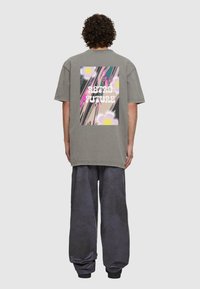 Pelēka pārsteidzīga t-krekls ar krāsainu "Retro nākotne" grafiku mugurā. Apģērbam ir īsas piedurknes un brīva piegriezuma siluets.